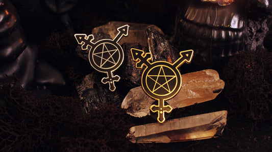 Two enamel pins, silver and gold, combining the trans symbol with a pentacle, displayed on smoky quartz crystals against a dark background