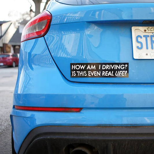 Back of blue car with black bumper sticker with white text "HOW AM I DRIVING? IS THIS EVEN REAL LIFE?" - PABOOM