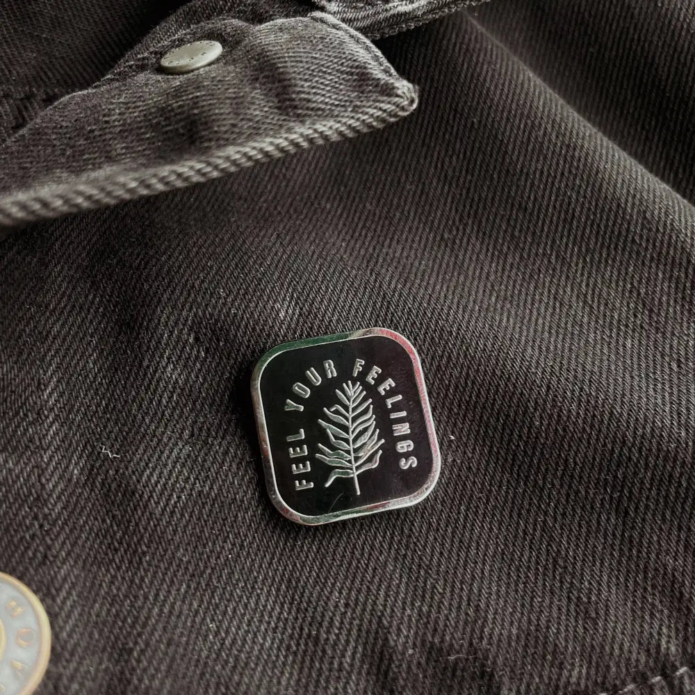 Black and silver lapel pin with a fern and text 'Feel your feelings" on grey fabric