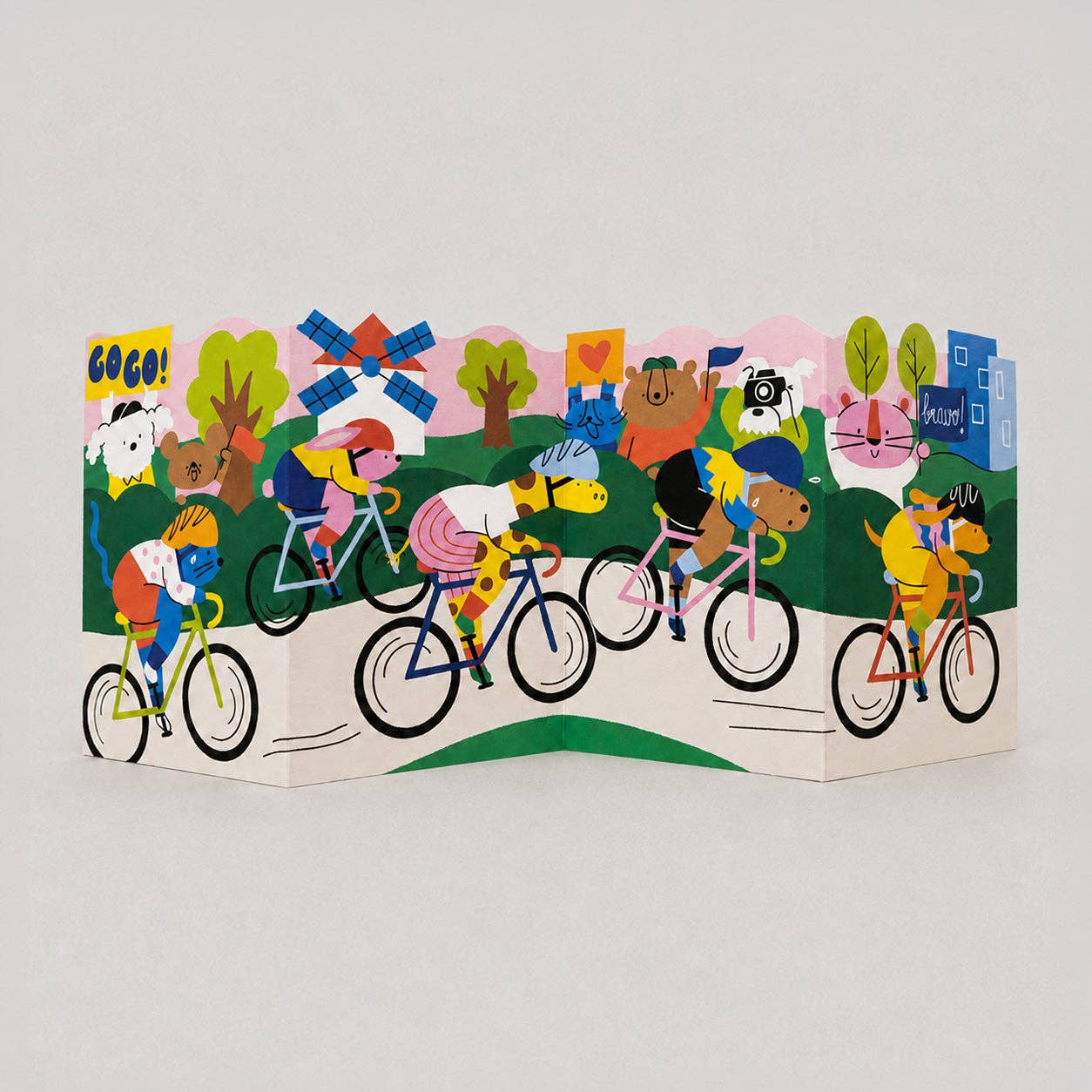 Illustrated concertina fold-out card unfolded to show a Tour de France-style cycling scene with animal characters riding bikes past a windmill and city buildings.