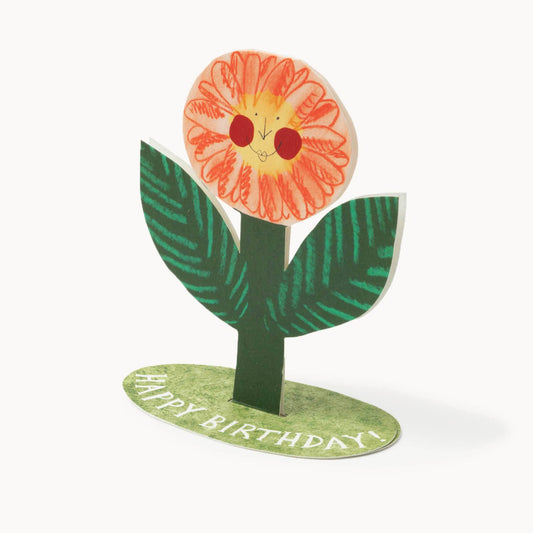 Free-standing hand-drawn orange flower with a smiling rosy-cheeked face, on a green grassy base reading 'Happy Birthday!'