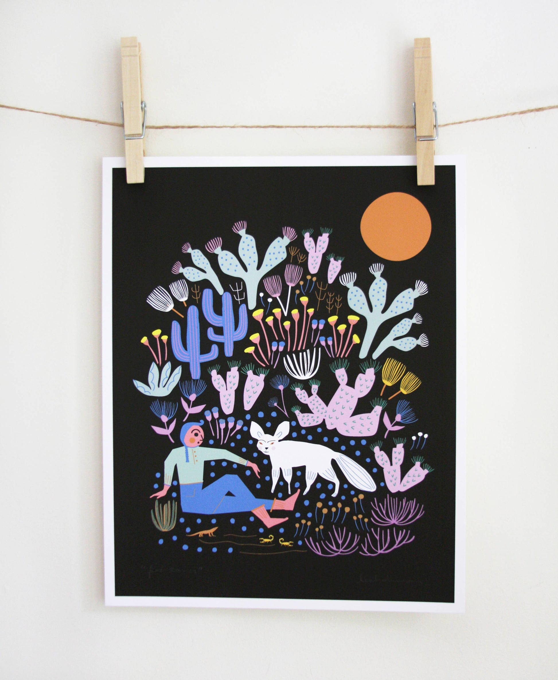 Art print of a person sitting with a white arctic fox among purple and blue cacti under an orange sun on a black background, clipped to a string — PABOOM