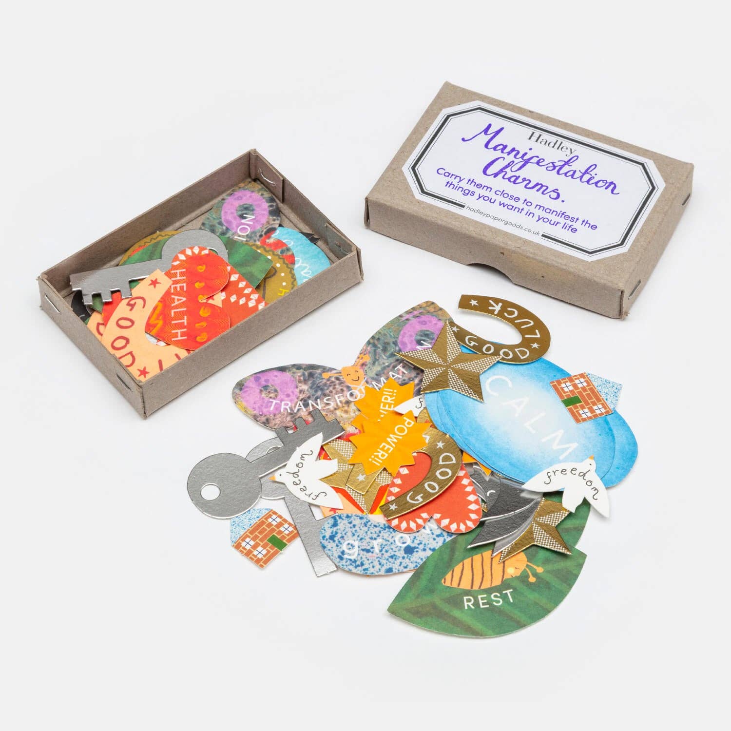 Open kraft box labeled 'Hadley Manifestation Charms, Carry Them Close to Manifest the Things You Want in Your Life' filled with colourful paper charms, with more charms fanned out in front reading Health, Good Luck, Calm, Transformation, Rest and Freedom