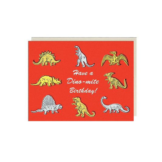 Red greeting card with eight illustrated dinosaurs in yellow, gray, and white surrounding silver foil script text "Have a Dino-mite Birthday!" - PABOOM