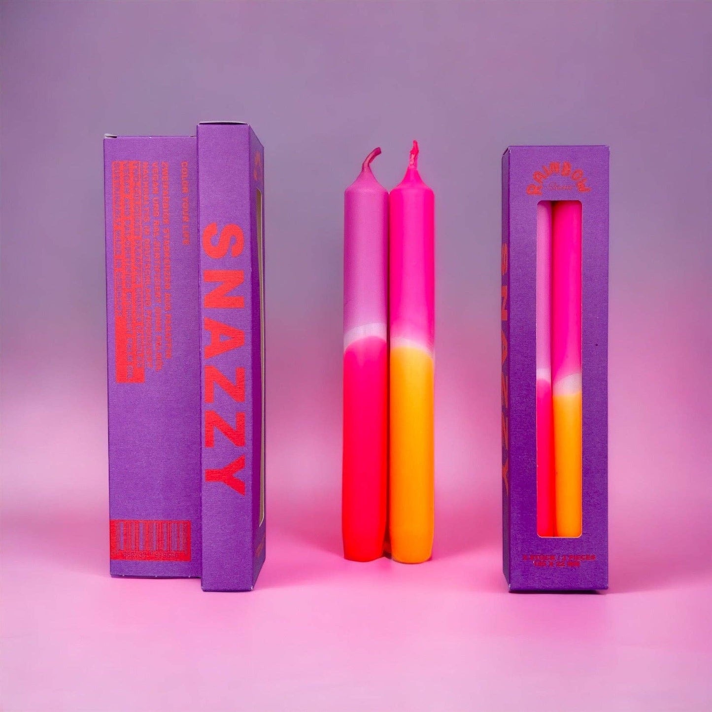 Rainbow Studio Dinner Candle Set Snazzy — two dip-dyed taper candles in pink-to-orange gradient, shown beside purple packaging