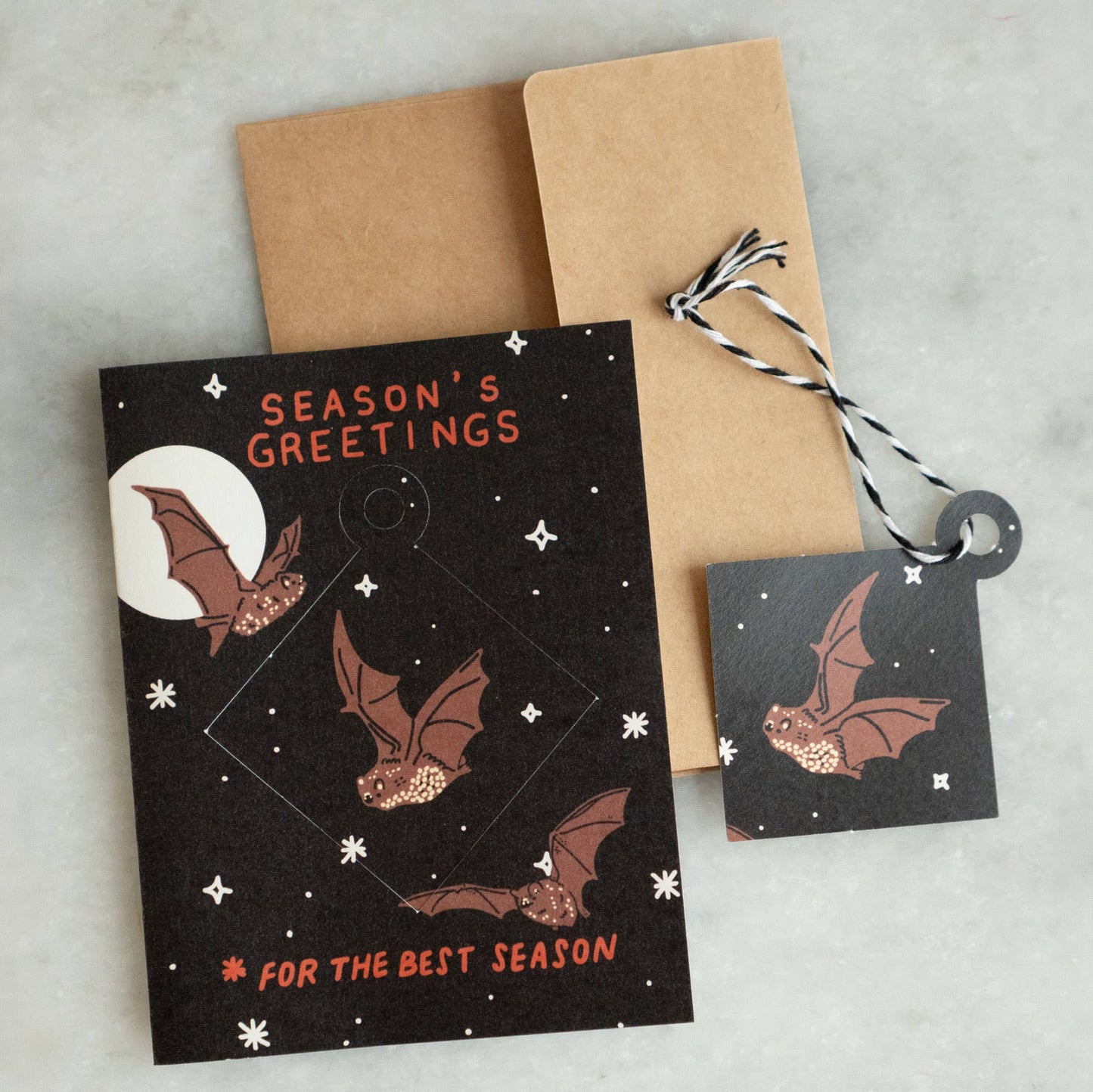 Black 'Season's Greetings, For the Best Season' greeting card with a punch-out diamond ornament featuring illustrated bats and a full moon, kraft envelope alongside
