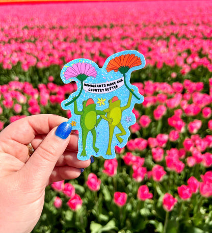 Two green frogs in party hats dancing and holding flowers on a glittery blue background, reading Immigrants make our country better