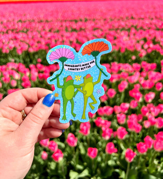 Two green frogs in party hats dancing and holding flowers on a glittery blue background, reading Immigrants make our country better