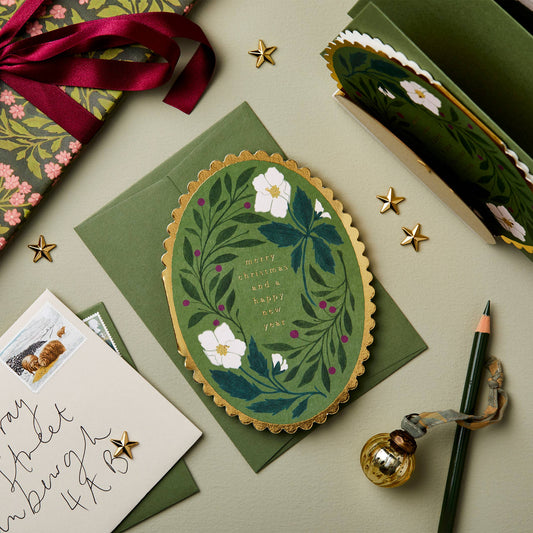 White hellebore flowers and berries on forest green oval card with gold scalloped edge, reading Merry Christmas and a Happy New Year