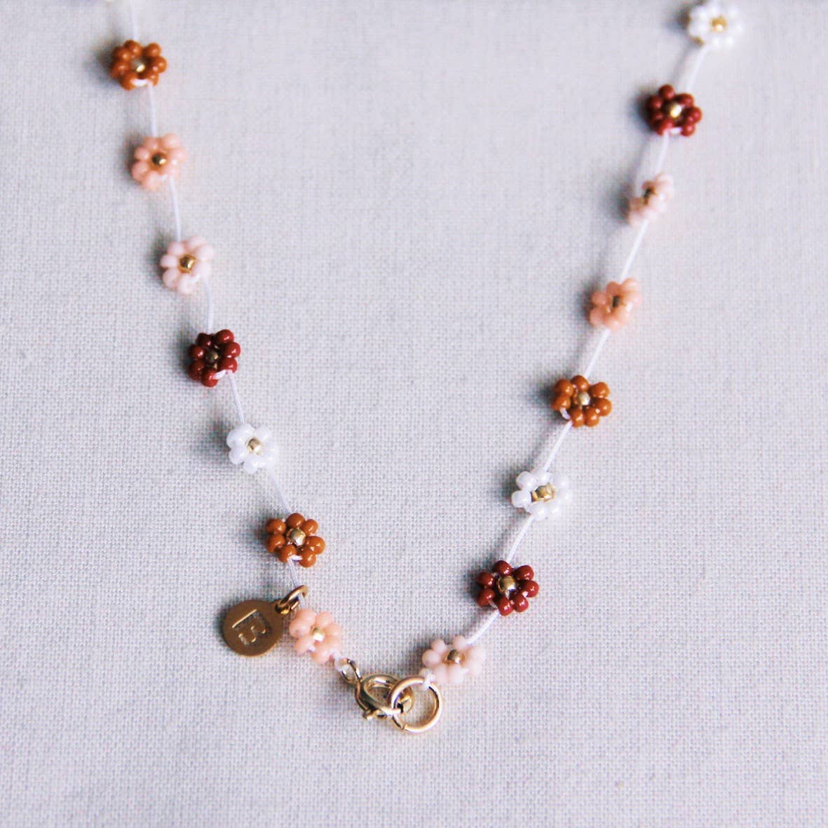 Handmade daisy flower beaded necklace in nude, blush, white and rust tones with a gold charm and lobster clasp, laid on linen fabric - PABOOM