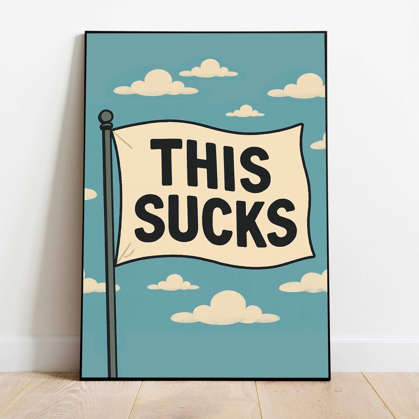 A framed art print leaning against a white wall on a light wood floor. The print features a retro-style illustration of a flag on a pole with the text 'THIS SUCKS' in bold black letters against a light blue sky with cream-colored clouds.