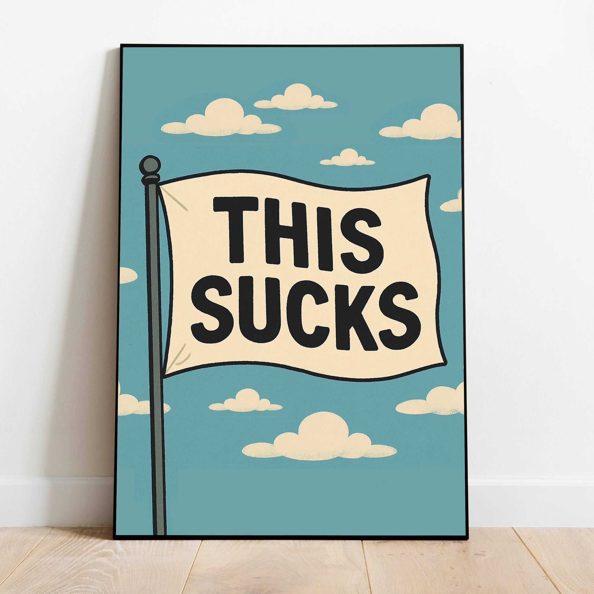 A framed art print leaning against a white wall on a light wood floor. The print features a retro-style illustration of a flag on a pole with the text 'THIS SUCKS' in bold black letters against a light blue sky with cream-colored clouds.