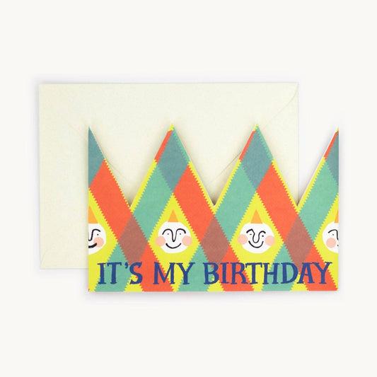 Multicoloured argyle-patterned party hat crown card with small smiling faces and the text 'IT'S MY BIRTHDAY' in navy, cream envelope behind