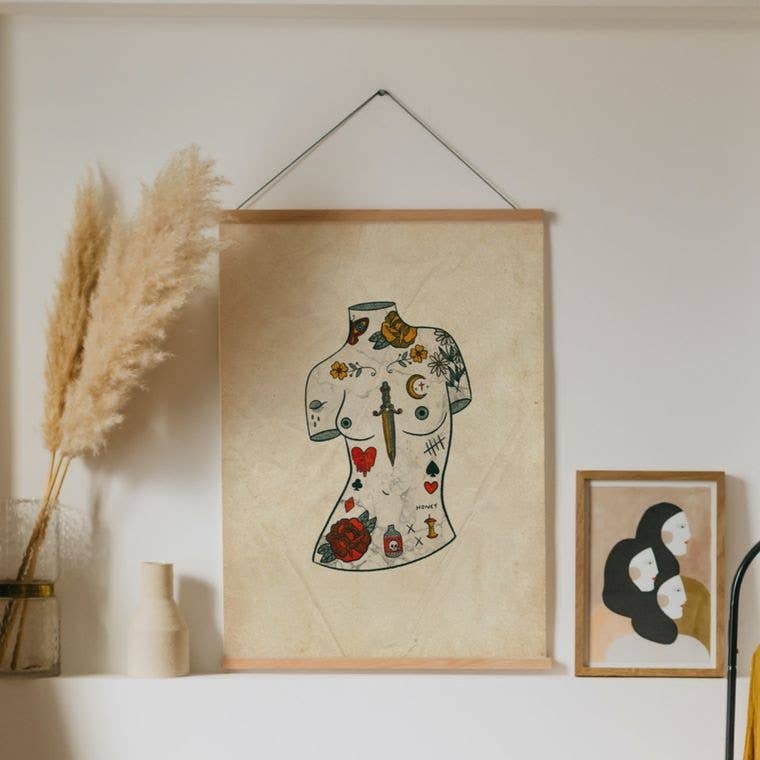 Framed art print of a tattooed classical statue figure in vintage pin-up tattoo style, displayed on a shelf with dried pampas grass