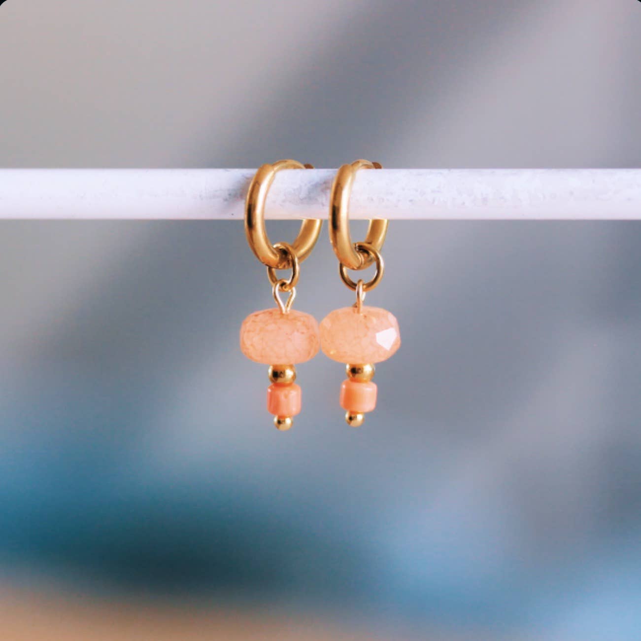 Gold-tone stainless steel huggie hoop earrings with a faceted coral stone bead dangle, shown hanging on a display bar - PABOOM