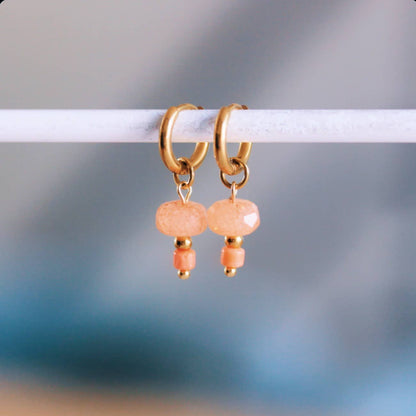 Gold-tone stainless steel huggie hoop earrings with a faceted coral stone bead dangle, shown hanging on a display bar - PABOOM