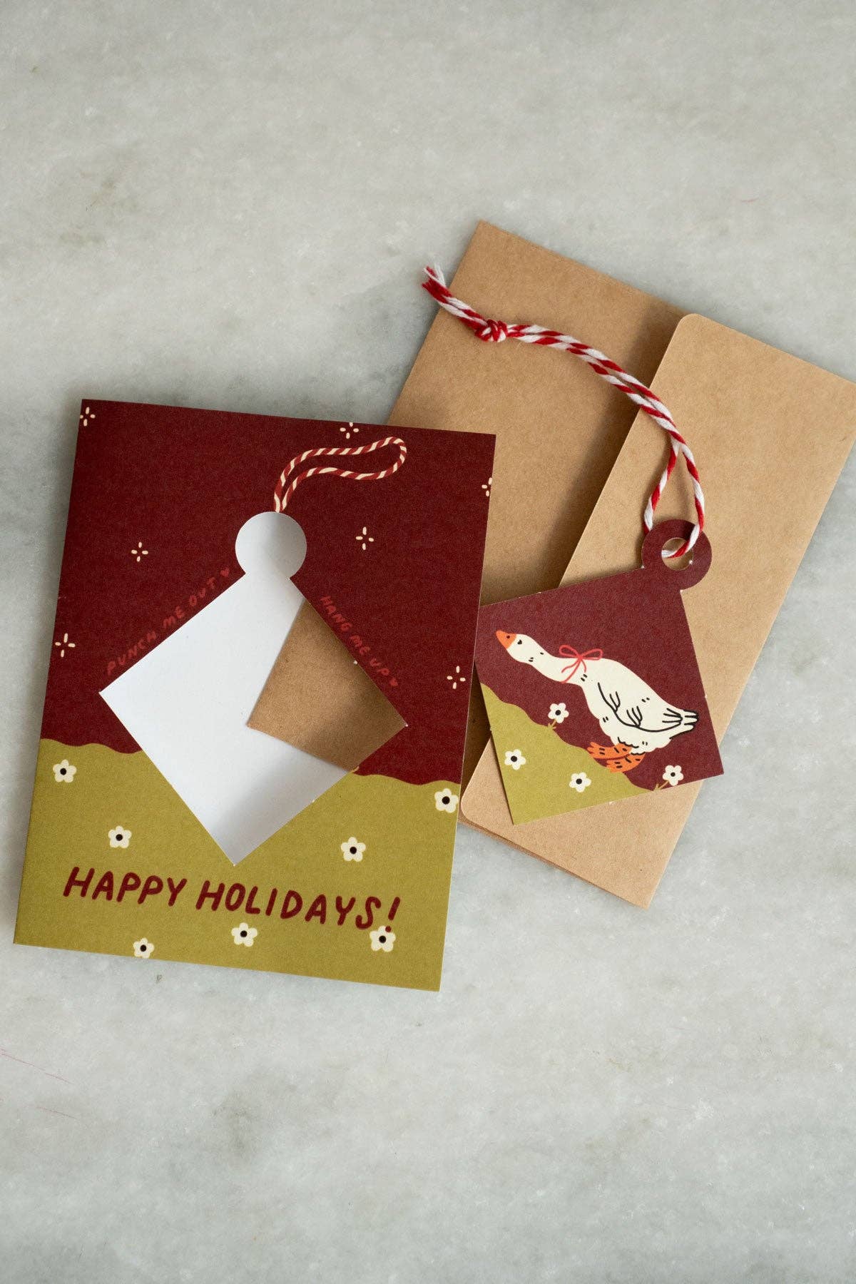 Maroon and olive green 'Happy Holidays' greeting card with a punch-out diamond ornament featuring an illustrated goose with a red bow, kraft envelope alongside
