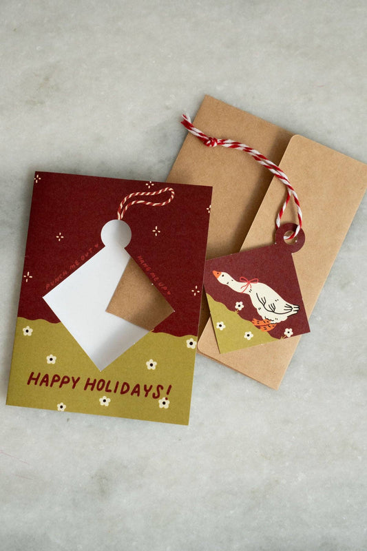 Maroon and olive green 'Happy Holidays' greeting card with a punch-out diamond ornament featuring an illustrated goose with a red bow, kraft envelope alongside