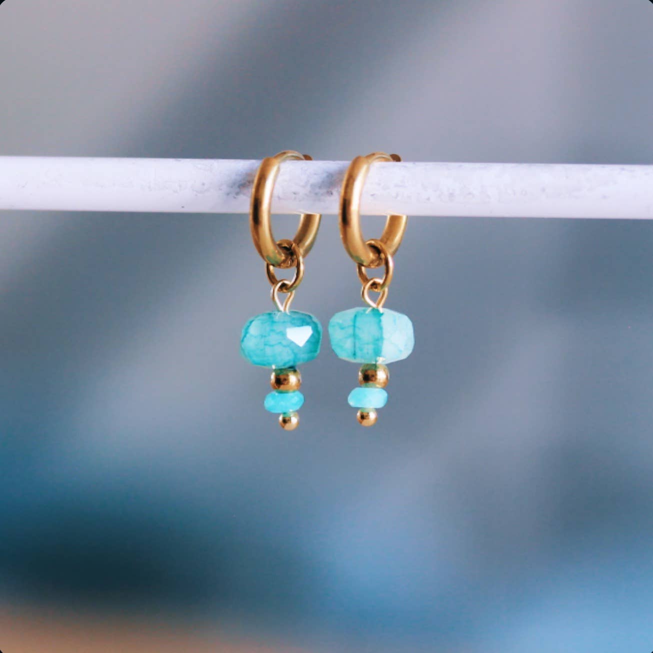 Gold-tone stainless steel huggie hoop earrings with a faceted sea green stone bead dangle, shown hanging on a display bar - PABOOM