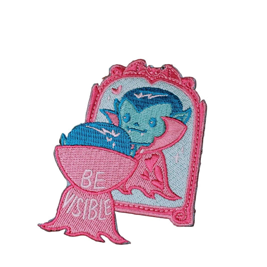 Pink and blue embroidered iron-on patch of a vampire bat character looking into an ornate mirror with the words Be Visible stitched below