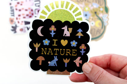 Black scalloped glitter vinyl sticker reading I Love Nature with fox, moon, mushroom, and flower icons, held in hand — PABOOM