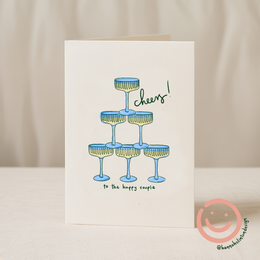 Blue Iris Designs Co wedding card illustrated with a stacked pyramid of blue champagne coupes and "cheers! to the happy couple" text - PABOOM