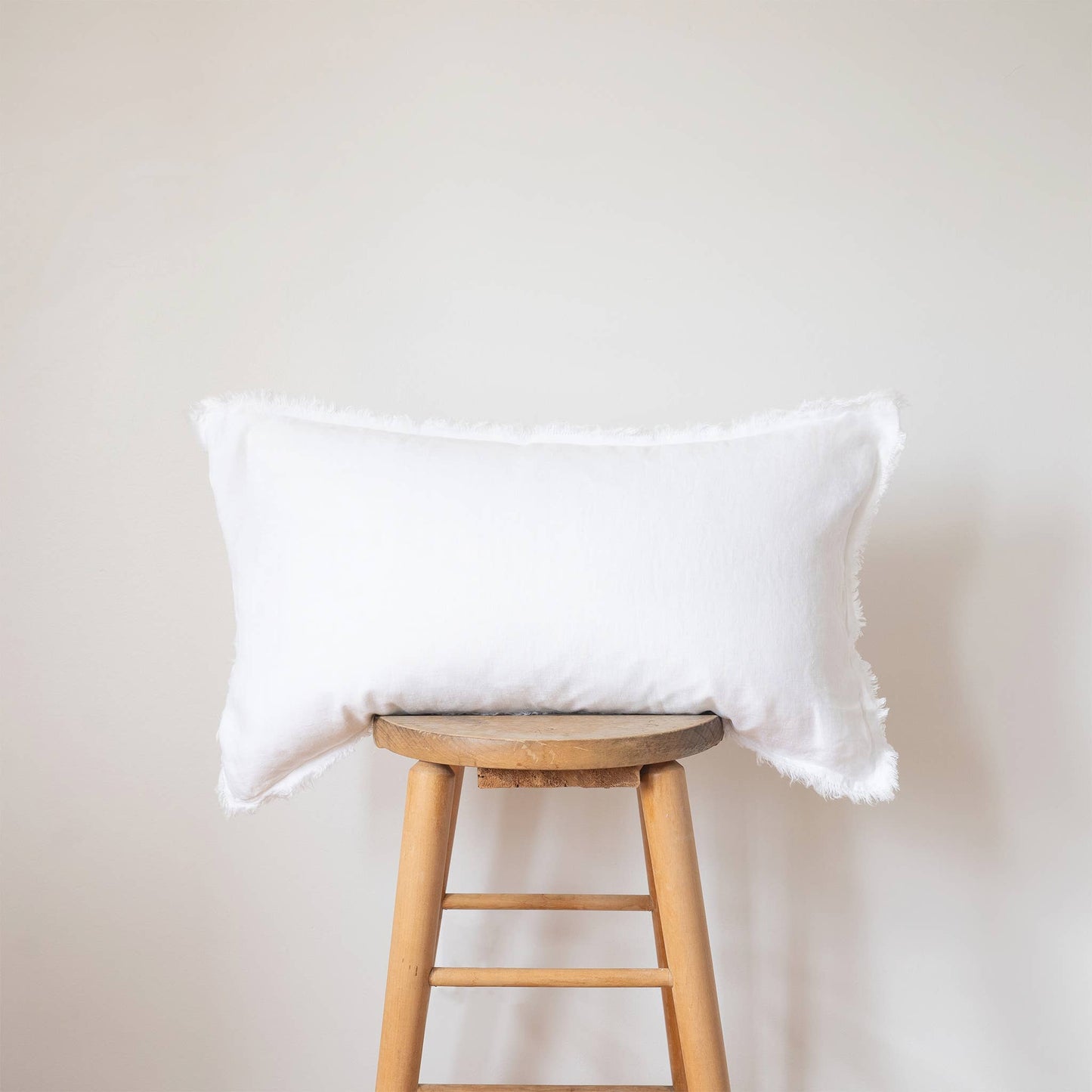 White linen lumbar pillow cover with a frayed fringe edge displayed on a wooden stool against a white wall