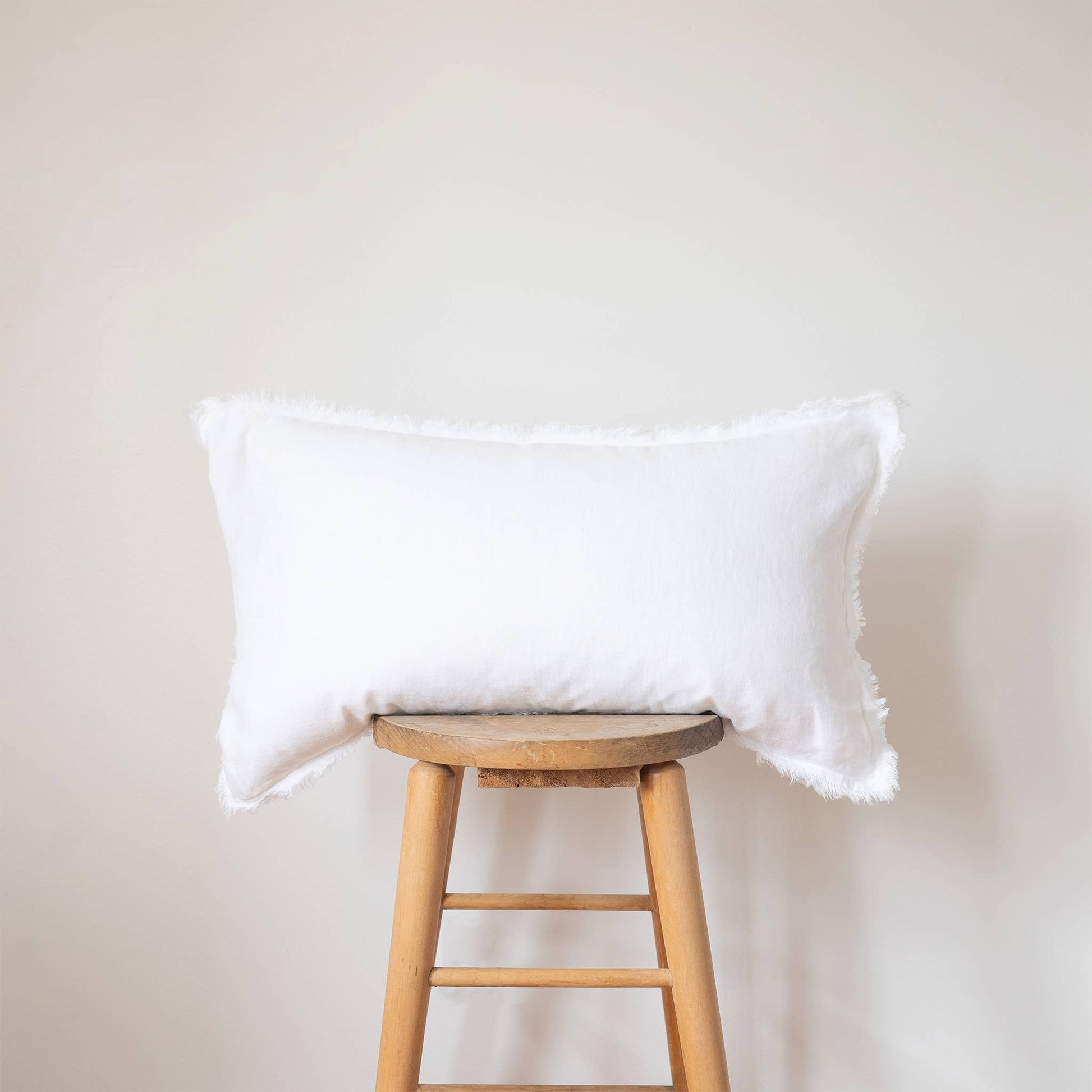 White linen lumbar pillow cover with a frayed fringe edge displayed on a wooden stool against a white wall