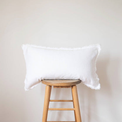 White linen lumbar pillow cover with a frayed fringe edge displayed on a wooden stool against a white wall