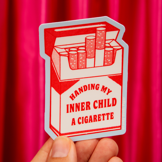 Matte vinyl sticker of a red and white illustrated cigarette pack with text "Handing My Inner Child A Cigarette," held up against a pink curtain background - PABOOM