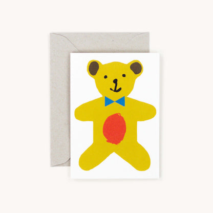 White greeting card featuring an illustrated yellow teddy bear with a blue bow tie and a red patch on its belly, grey envelope behind
