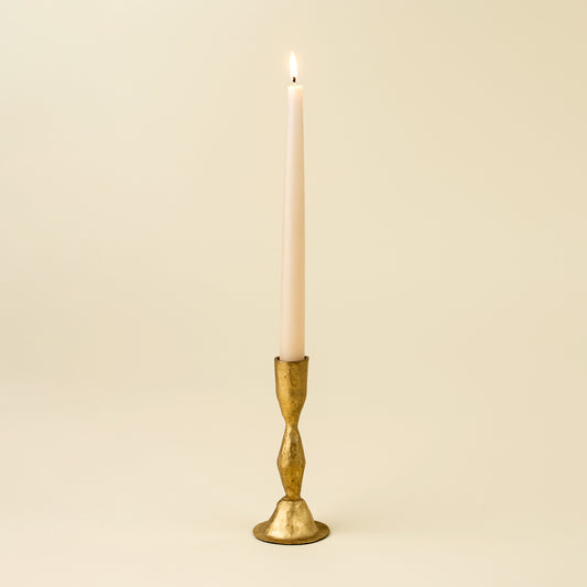 Tall gold faceted candlestick holding a lit taper candle against a cream background
