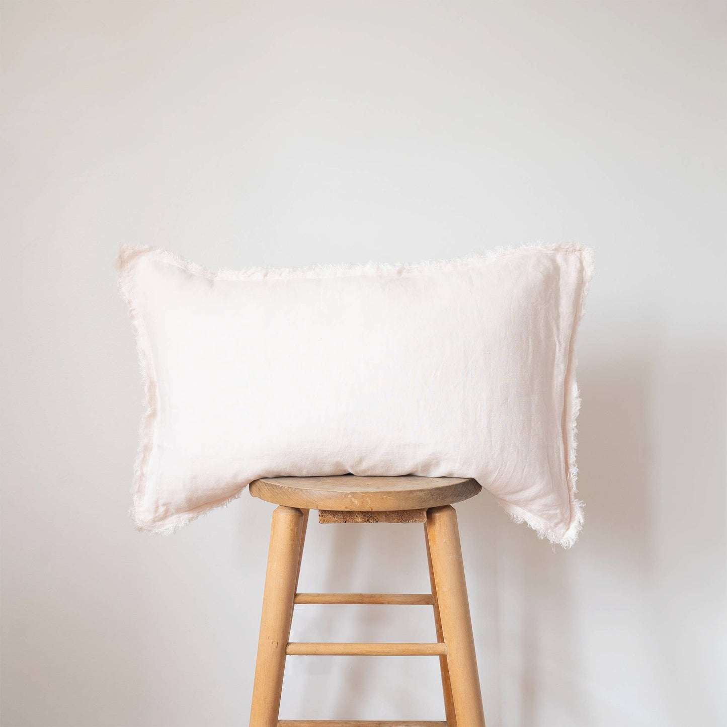 Blush pink linen lumbar pillow cover with a frayed fringe edge displayed on a wooden stool against a white wall