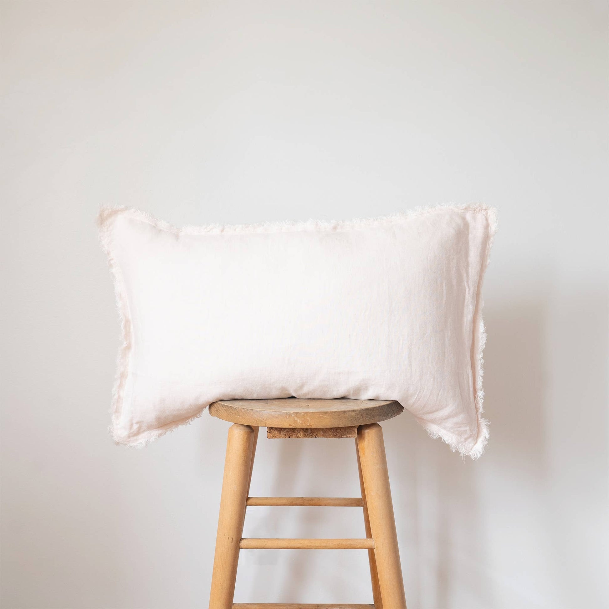 Blush pink linen lumbar pillow cover with a frayed fringe edge displayed on a wooden stool against a white wall