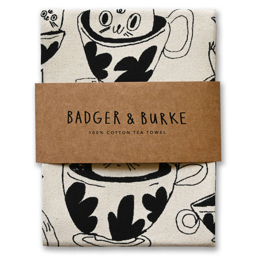 Folded natural cotton tea towel printed with black teacups and a cat peeking out, Badger + Burke kraft band - PABOOM