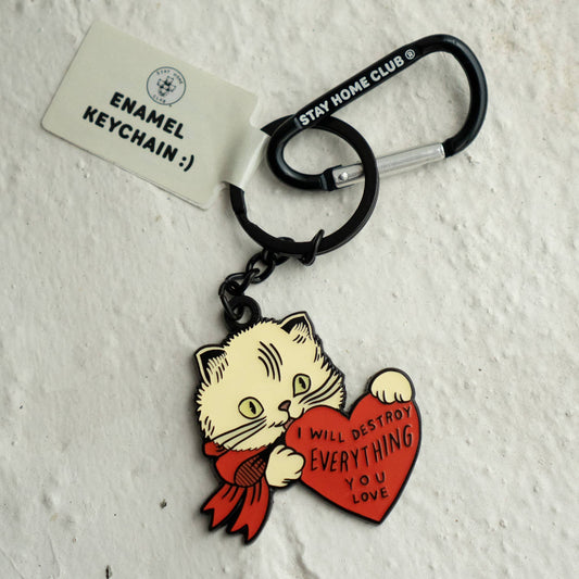 Black enamel cat keychain holding a red heart that reads 'I will destroy everything you love,' attached to a Stay Home Club branded carabiner on a textured white background