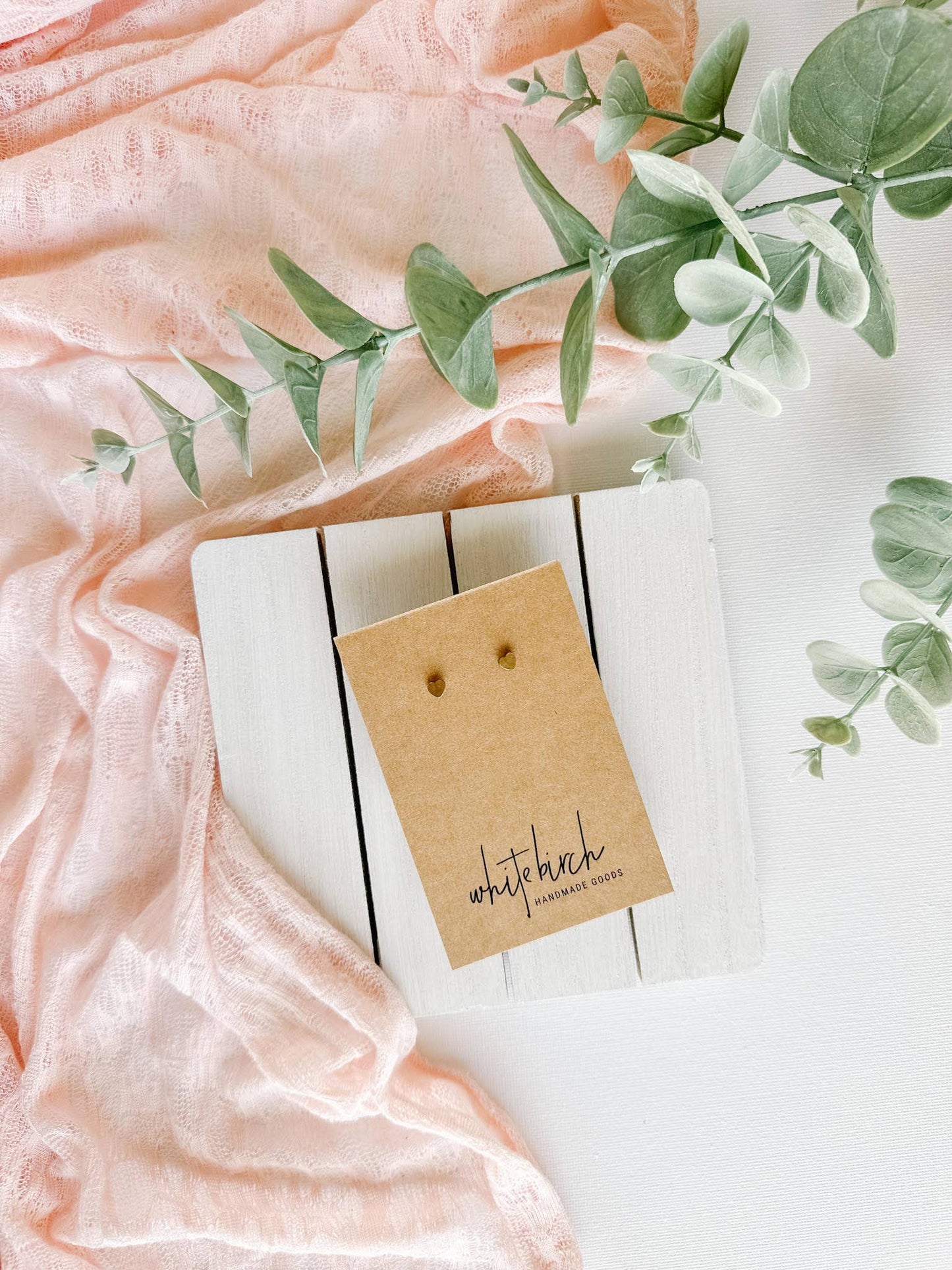 Mini raw brass heart stud earrings on a Whitebirch Handmade Goods earring card with a blush scarf and eucalyptus