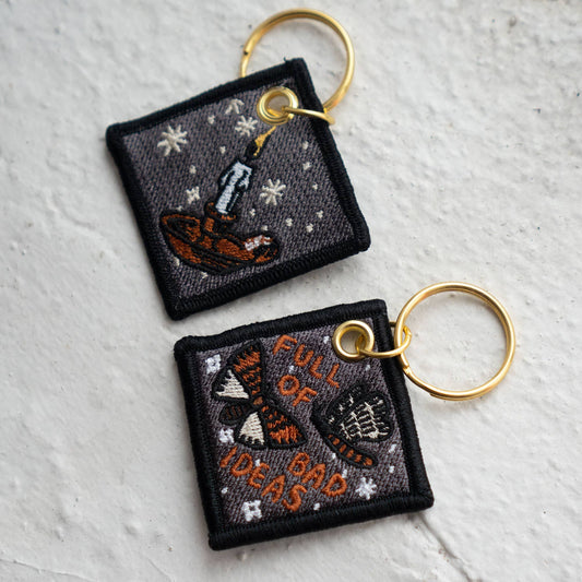 Two square embroidered keychains on gold-tone rings — one shows a lit candle with sparkles, the other reads 'Full of Bad Ideas' with two moths, on a textured white wall