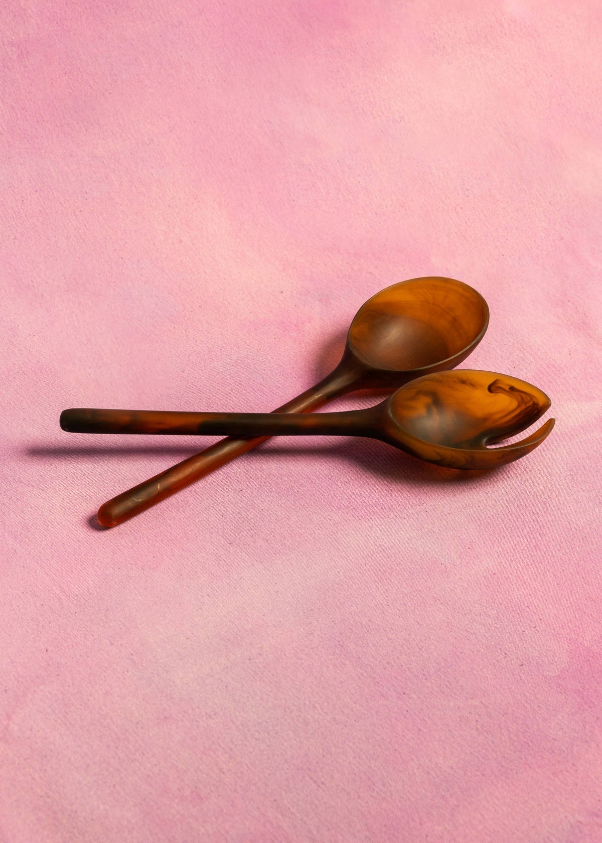 Marbled amber resin salad server fork and spoon crossed on a soft pink textured backdrop