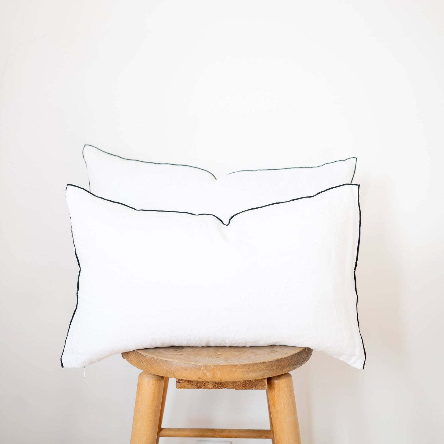 Two white linen lumbar pillow covers with black contrast-edge trim in 12x20 and 14x22 sizes stacked on a wooden stool against a white wall