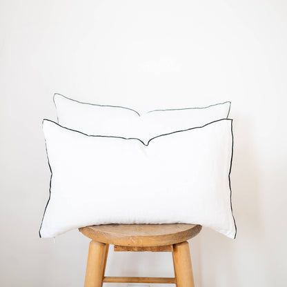 Two white linen lumbar pillow covers with black contrast-edge trim in 12x20 and 14x22 sizes stacked on a wooden stool against a white wall