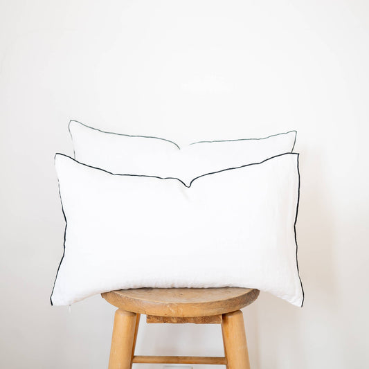 Two white linen lumbar pillow covers with black contrast-edge trim in 12x20 and 14x22 sizes stacked on a wooden stool against a white wall