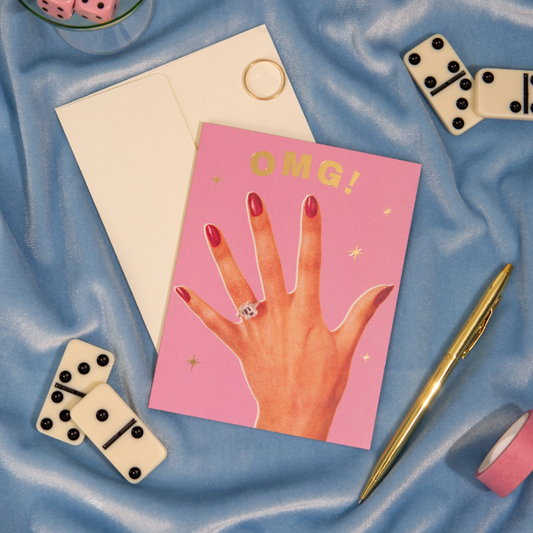 Pink greeting card with gold foil text "OMG!" above an illustrated hand with red manicured nails wearing an engagement ring, gold foil sparkle accents - PABOOM