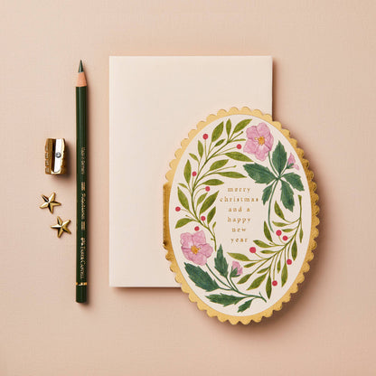 Pink floral wreath with red berries on oval card with gold scalloped edge, reading Merry Christmas and a Happy New Year
