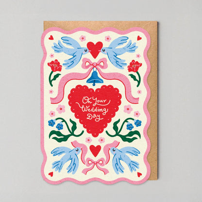 Scalloped-edge wedding greeting card with pink border, illustrated lovebirds, a ribbon-tied bell, and a red heart reading "On Your Wedding Day."