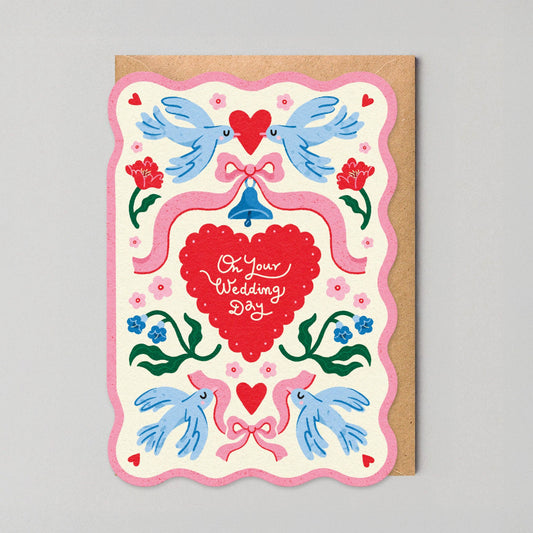 Scalloped-edge wedding greeting card with pink border, illustrated lovebirds, a ribbon-tied bell, and a red heart reading "On Your Wedding Day."