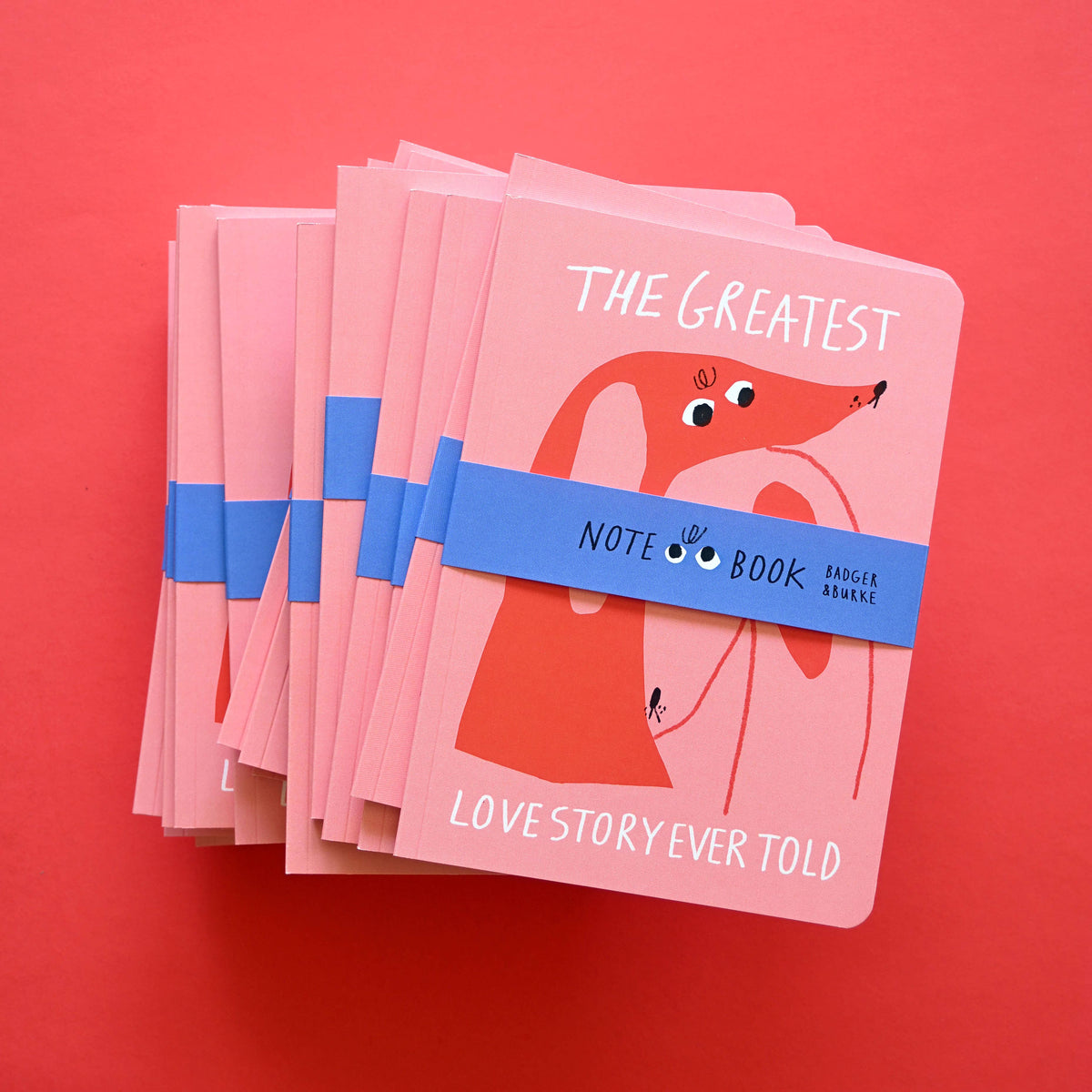Stack of pink The Greatest Love Story Ever Told notebooks with a red illustrated dog and blue Badger + Burke band - PABOOM
