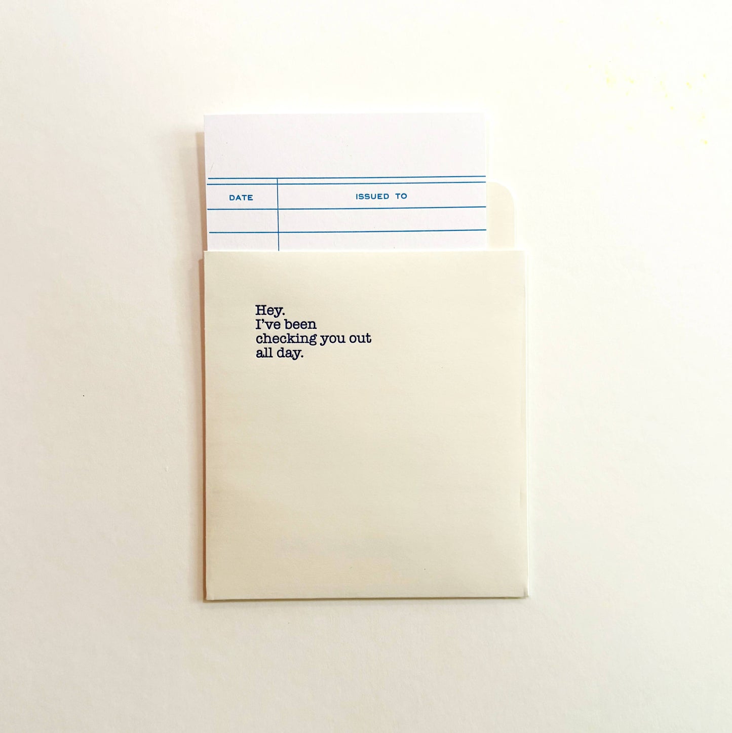 Flat lay of the light ivory library-card pocket and checkout insert, both printed with Hey, I've been checking you out all day