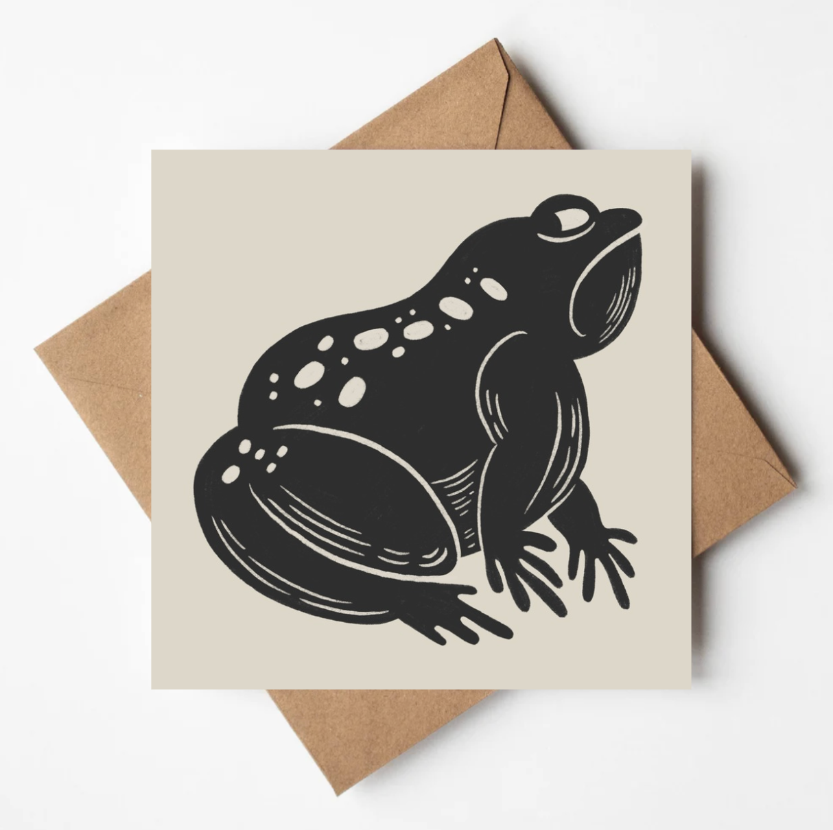 'The Grumpy Frog' animal card featuring a bold black illustration of a scowling toad on a cream square card, propped on a kraft envelope