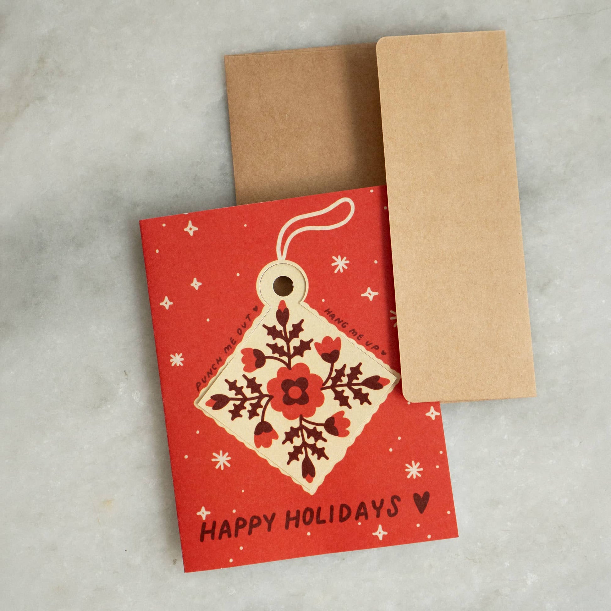 Red 'Happy Holidays' greeting card with a punch-out diamond ornament featuring a quilt-inspired floral pattern, kraft envelope alongside